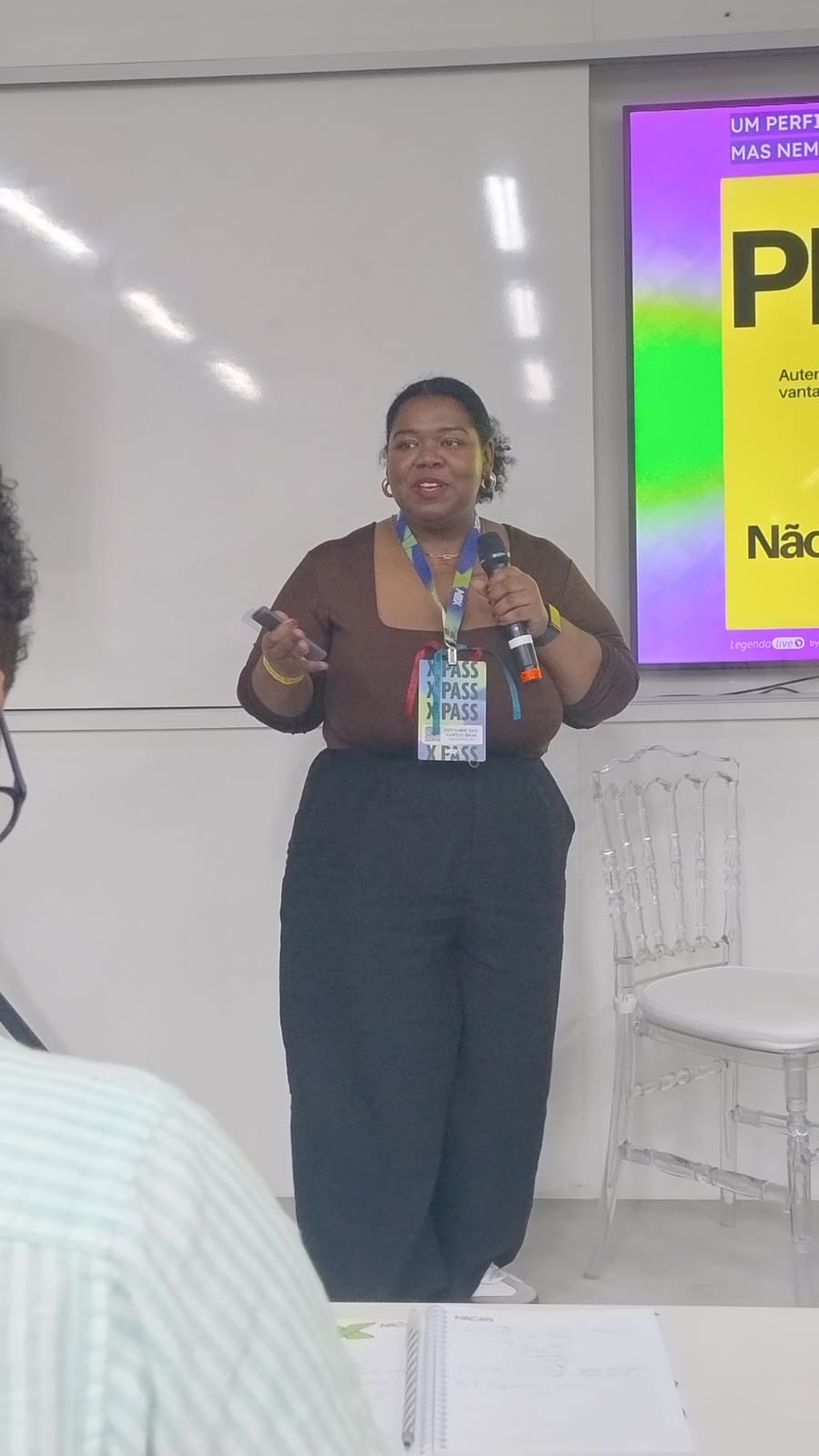 Ketyanne em pé, com microfone e passador de slides, apresentando para uma turma