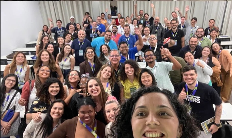 Selfie de grupo com dezenas de participantes sorrindo e acenando em uma sala de workshop